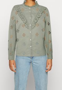 Lichtgroen overhemd met knopen en kantafwerking, met uitsparings en gekleurde accenten in oranje, blauw en zwart. Lange mouwen en een ontspannen pasvorm.