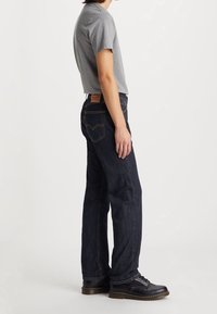 Vaqueros de mezclilla azul oscuro con un ajuste entallado, que presentan una parcha de cuero en la cintura y detalles cosidos cerca de los bolsillos. Combinados con una camiseta gris y zapatos negros.