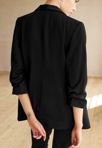 Personne en blazer noir avec les manches retroussées jusqu'aux coudes, montrant un tatouage de fleur de lotus sur le poignet gauche, debout sur un sol en bois clair.