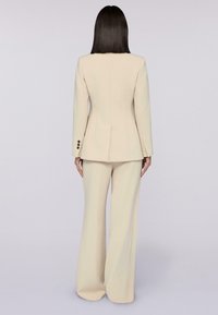 Crème-kleurig op maat gemaakt pak met een gestructureerde blazer met een enkele split en uitlopende broekspijpen. Zachte textuur en slanke silhouette.