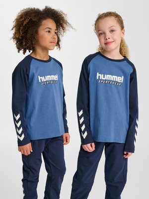 Deux enfants portant des vêtements de sport Hummel bleu et marine avec des motifs chevrons sur les manches et les pantalons, debout devant un fond blanc uni.