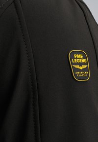 Zwarte jas met zichtbare stiksels en een geel en zwart PME LEGEND-patch. Soepele textuur met een eigentijds ontwerp.