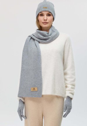 Cappello di lana a coste grigio e sciarpa abbinata con guanti grigi. Maglione soffice di colore chiaro e pantaloni beige. Logo in pelle sulla sciarpa.