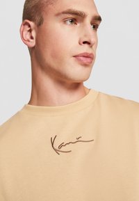 Beige strikkegenser med ribbestrikket hals, med mørkebrun brodert signaturdetalj foran. Glatt tekstur, avslappet passform.