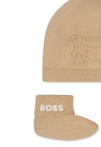 Bonnet en tricot beige avec un motif animal en relief ; chaussons assortis avec "BOSS" en lettres blanches sur le bord. Texture douce, forme arrondie.