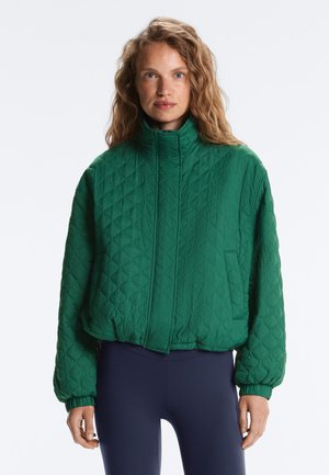 Femeie cu păr creț purtând o jachetă verde matlasată cu guler înalt și colanți bleumarin, stând în fața unui fundal simplu.