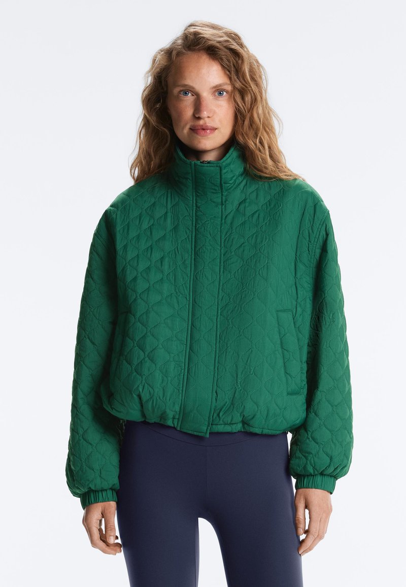 Femme aux cheveux bouclés portant une veste matelassée verte à col haut et des leggings bleu marine, debout devant un fond uni.