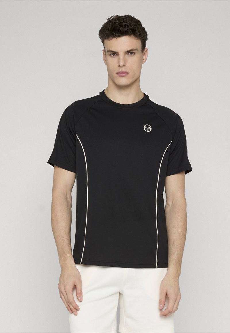Jeune homme portant une chemise noire à manches courtes avec un liseré blanc et un logo blanc sur la poitrine, assortie à un pantalon blanc, debout devant un fond gris.