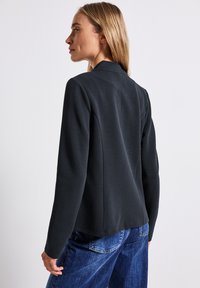 Svart texturerad kavaj med krage, långa ärmar och öppen front. Bärs över blå denimbyxor.