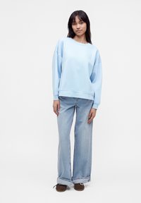 Jeune femme portant un sweat-shirt oversize bleu clair, un jean bleu clair à jambes larges avec revers roulés et des chaussures marron, debout sur fond blanc.