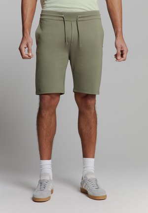Olijfgroen katoenen shorts met een elastische tailleband en trekkoord. Beschikt over zijzakken en een gladde textuur, knielang model, gedragen met grijze sneakers.