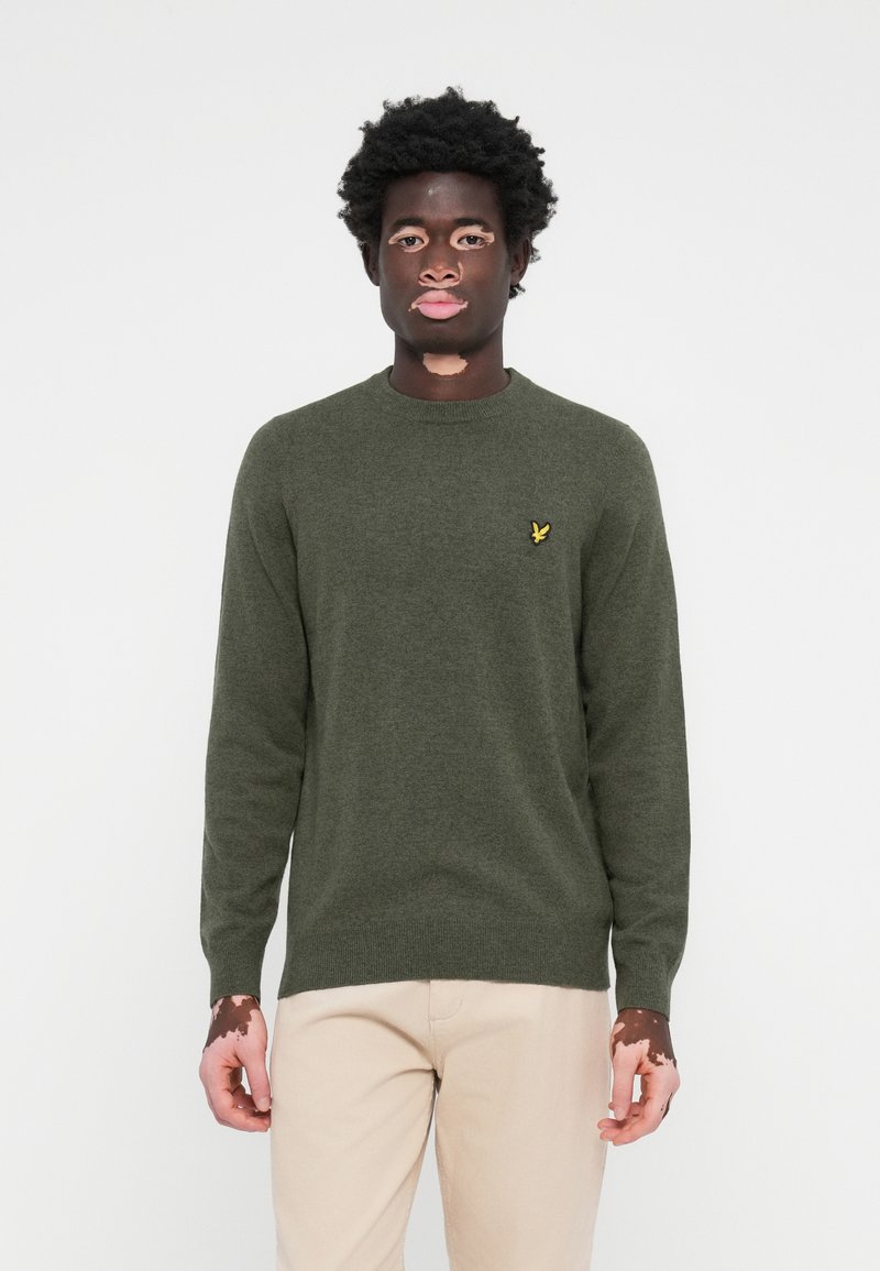 Pull en tricot vert avec un col rond et des manches longues. Présente un petit logo jaune sur la poitrine. Texture douce et coupe ajustée.