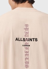 Camiseta de algodón beige con un logo vertical en negro "ALLSAINTS LONDON" y texto bordado en rosa debajo. Diseño clásico de cuello redondo.