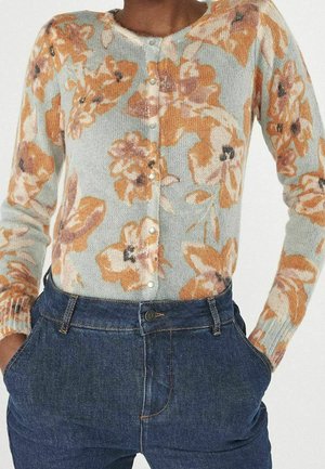 Frau trägt eine hellblaue, geknöpfte Strickjacke mit großen orangefarbenen Blumenmustern und dunkle, hoch taillierte Jeans, Hände in den Taschen.