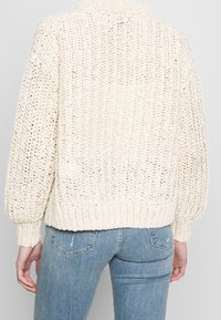 Cremefarbener gestrickter Pullover mit lockerer Passform, einer gerippten Saum- und Manschettenstruktur sowie einer offen gewebten Textur.