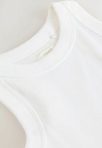 Vit ribbad tanktop med rund halsringning och sydda detaljer. Tyget verkar slätt med lätt struktur, märkt "NEXT."