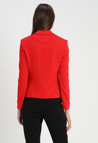 Blazer rouge sur mesure avec une silhouette ajustée, des épaules structurées et un tissu lisse. Comprend un col classique et des détails de coutures arrière.