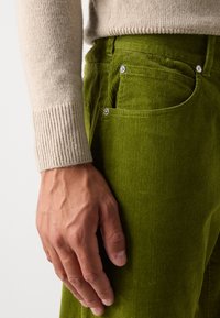Grønne fløjlsbukser med struktureret stof, synlige søm detaljer og blankt sølv hardware. Beigefarvet strikket sweaterskæftet er delvist synligt.