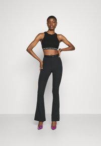 Zwarte cropped tanktop met logo-band, gecombineerd met aansluitende zwarte flared broeken. Model draagt roze en paarse slangenprint-hakken.