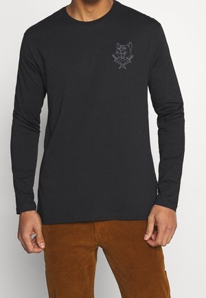 Mann trägt ein schwarzes Langarmshirt mit einem kleinen wütenden Katzen- und gekreuzte Messer-Motiv auf der Brust, kombiniert mit braunen Cordhosen.