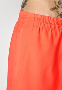 Nærbilde av lyse oransje elastiske shorts med sidelomme og delvis tatovering synlig på bærerens liv.