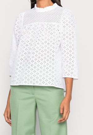 Vrouw draagt een witte blouse van oogkant met driekwart mouwen en een gerimpelde halslijn, gecombineerd met lichtgroene broek.