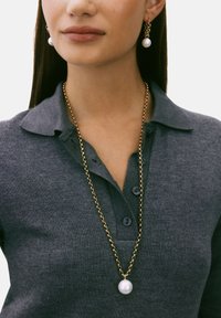 Zlatna ogrlica s velikom bijelom bisernom privjeskom, u kombinaciji s usklađenim bisernim visećim naušnicama. Model nosi tamnosivu polo majicu.
