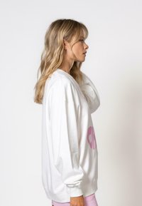 Sweatshirt oversize blanc avec du texte rose sur le devant, grandes manches longues, poignets côtelés et un tissu doux et texturé.