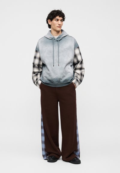 Νέος άνδρας με σγουρά μαλλιά που φοράει γκρι φούτερ με κουκούλα και καρό μανίκια, καφέ φαρδιά παντελόνια με καρό πλαϊνές ρίγες, και μαύρα παπούτσια.