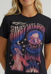 Camiseta de algodón negra con un diseño gráfico colorido de una mujer, estrellas y detalles de texto. Los colores del texto transicionan de rojo a azul. Mangas cortas.