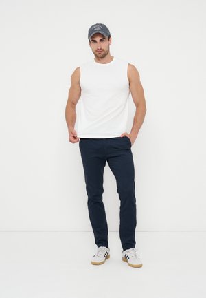 Fehér ujjatlan pólót, tengerészkék nadrágot, fehér tornacipőt és sötétszürke sapkát viselő férfi, sima fehér háttér előtt áll.