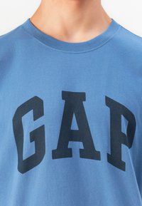Prim-plan al unei persoane îmbrăcate cu un tricou albastru cu guler rotund, având litere mari de culoare albastru închis care formează cuvântul "GAP" pe piept.