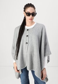 Poncho cinza de malha com colarinho arredondado, apresentando três botões pretos e costura branca em contraste nas bordas. Usado por cima de uma camisa branca.