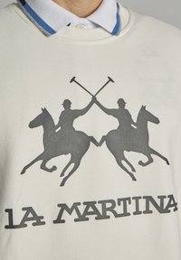 Pulover v odtenku off-white, ki ima velik sivi grafični motiv dveh polo igralcev na konjih, z besedilom "LA MARTINA" spodaj.