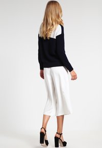 Navy- und weißfarbener, farbblockierter Pullover mit langen Ärmeln, kombiniert mit weißen, weiten Culottes und schwarzen, hohen Sandalen. Insgesamt glatte Textur.