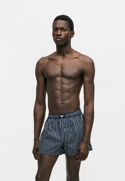 Μυώδης άνδρας με σορτσάκι μποξεράκι σε ναυτικό μπλε και λευκές κατακόρυφες ρίγες, στέκεται μπροστά σε απλό φωτεινό φόντο.