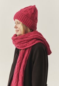 Roze gebreide muts met gestructureerd patroon en bijpassende dikke sjaal, beide met een kabelbreipatroon, gedragen over een donkere jas.