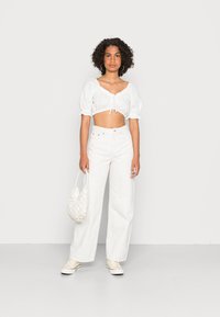 Blusa curta branca com mangas bufantes e detalhe de laço na frente, combinada com calças brancas de perna larga de cintura alta. Segura uma bolsa tecida branca.