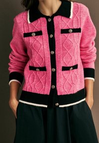 Femme portant un cardigan en maille torsadée rose avec bordure noire, boutons dorés et jupe noire, les mains dans les poches.