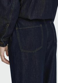 Jeans de mezclilla azul oscuro con un corte clásico, que presentan un bolsillo trasero con costuras amarillas y una cinturilla de altura media. Textura suave.