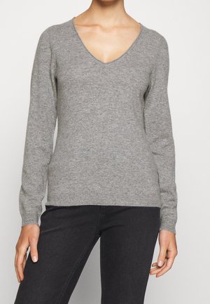 Personne portant un pull gris à manches longues en V et un pantalon noir, debout devant un fond blanc uni.