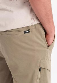 Shorts cargo beiges avec une poche à rabat, un détail zippé et une étiquette en tissu. Le matériau semble solide avec une texture lisse.