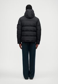 Jachetă puffere neagră cu glugă, având un design matlasat, umplutură groasă și mâneci lungi, asociată cu blugi largi de culoare albastru închis.