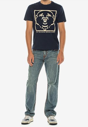 Mand iført mørk grafisk t-shirt med Buddhaansigt og tekst, lyseblå jeans og hvide sneakers, der står mod en hvid baggrund.
