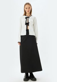 Witte gebreide cardigan met zwarte accenten, open voorkant en een striksluiting; gecombineerd met een lange zwarte rok en stevige zwarte laarzen.