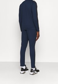Homme debout de dos portant un survêtement bleu marine avec des bandes latérales noires et des baskets noires et blanches avec des chaussettes blanches.