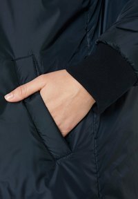 Gepolsterte, dunkelblaue Jacke mit seitlicher Tasche und gerippten schwarzen Strickbündchen. Glattes, glänzendes Außenmaterial mit subtiler Satinoberfläche.