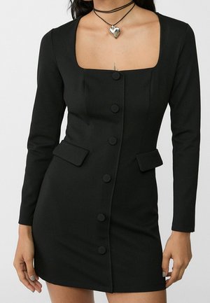 Kobieta ubrana w dopasowaną czarną sukienkę z długimi rękawami, kwadratowym dekoltem, guzikami z przodu, kieszeniami z klapkami oraz warstwowy naszyjnik z zawieszką w kształcie serca.
