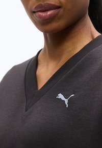 Κοντινό πλάνο ενός ατόμου που φοράει σκούρο γκρι μπλουζάκι με V λαιμόκοψη και μικρό λευκό λογότυπο Puma στο στήθος.