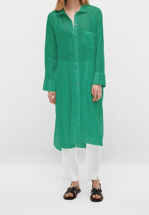 Osoba w długiej zielonej koszuli zapinanej na guziki z kieszenią na piersi, białych szerokich spodniach i czarnych sandałach na otwarte palce, stojąca na jednolitym tle.